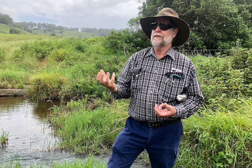 A man in a checked shirt gesticulates with his hands on the side of a creek.