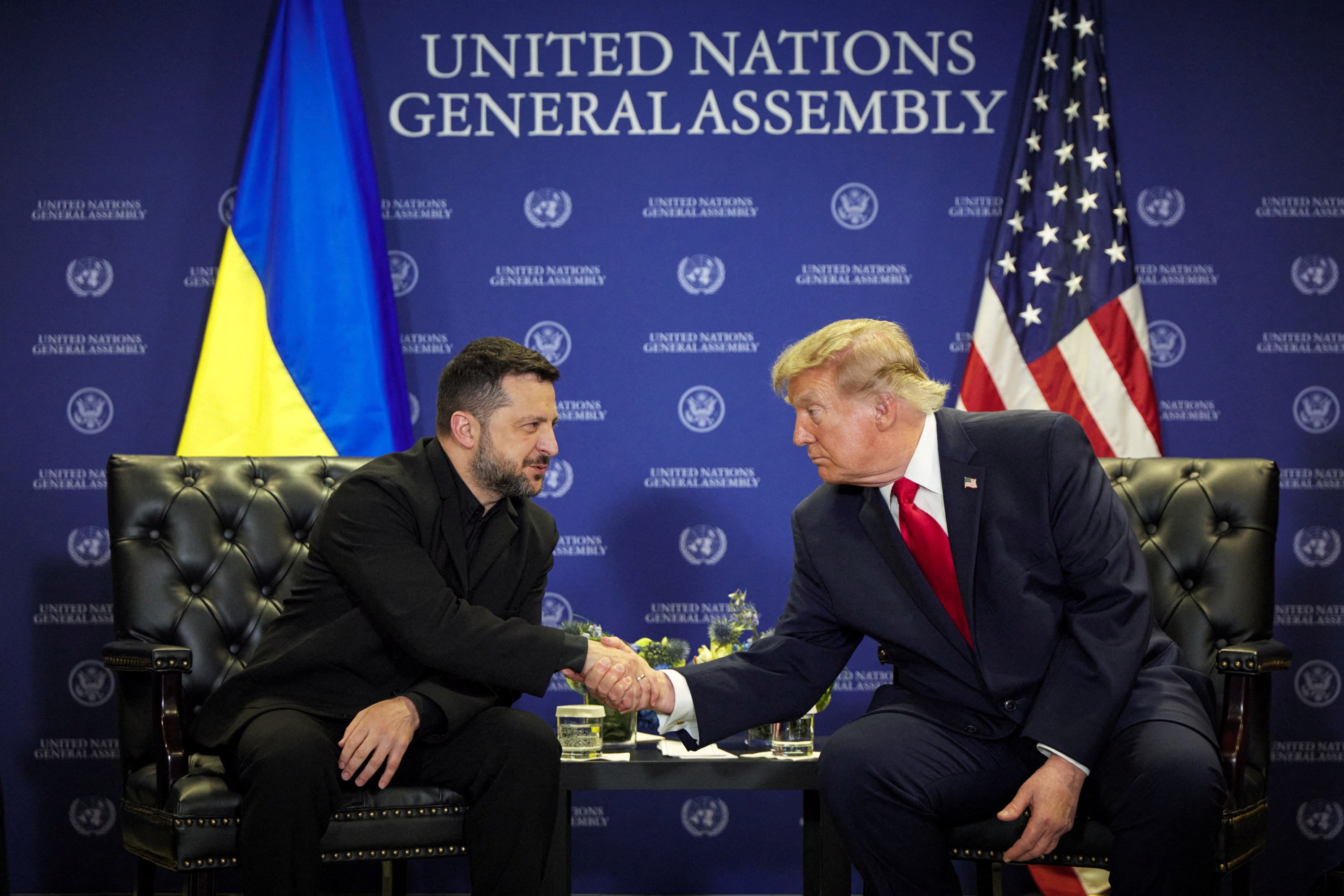 Volodymyr Zelenskyy and Donald Trump shake hands while seated in front of a UN wall.