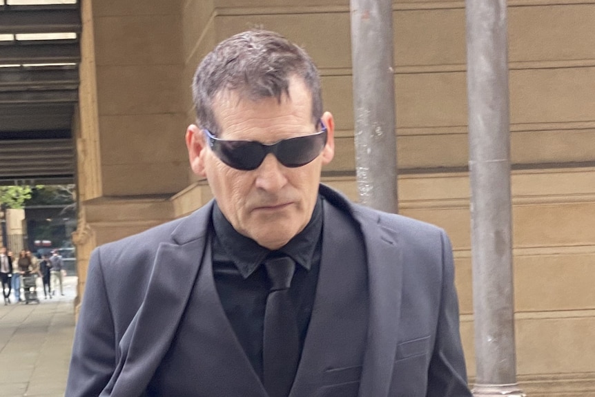 A man wearing a grey suit with black shirt and tie and sunglasses walks outside a court building with a serious expression