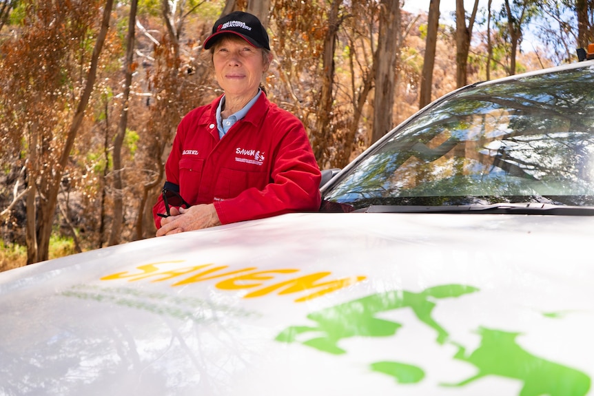 A woman earing red overalls and a cap stands behind a car bonnet