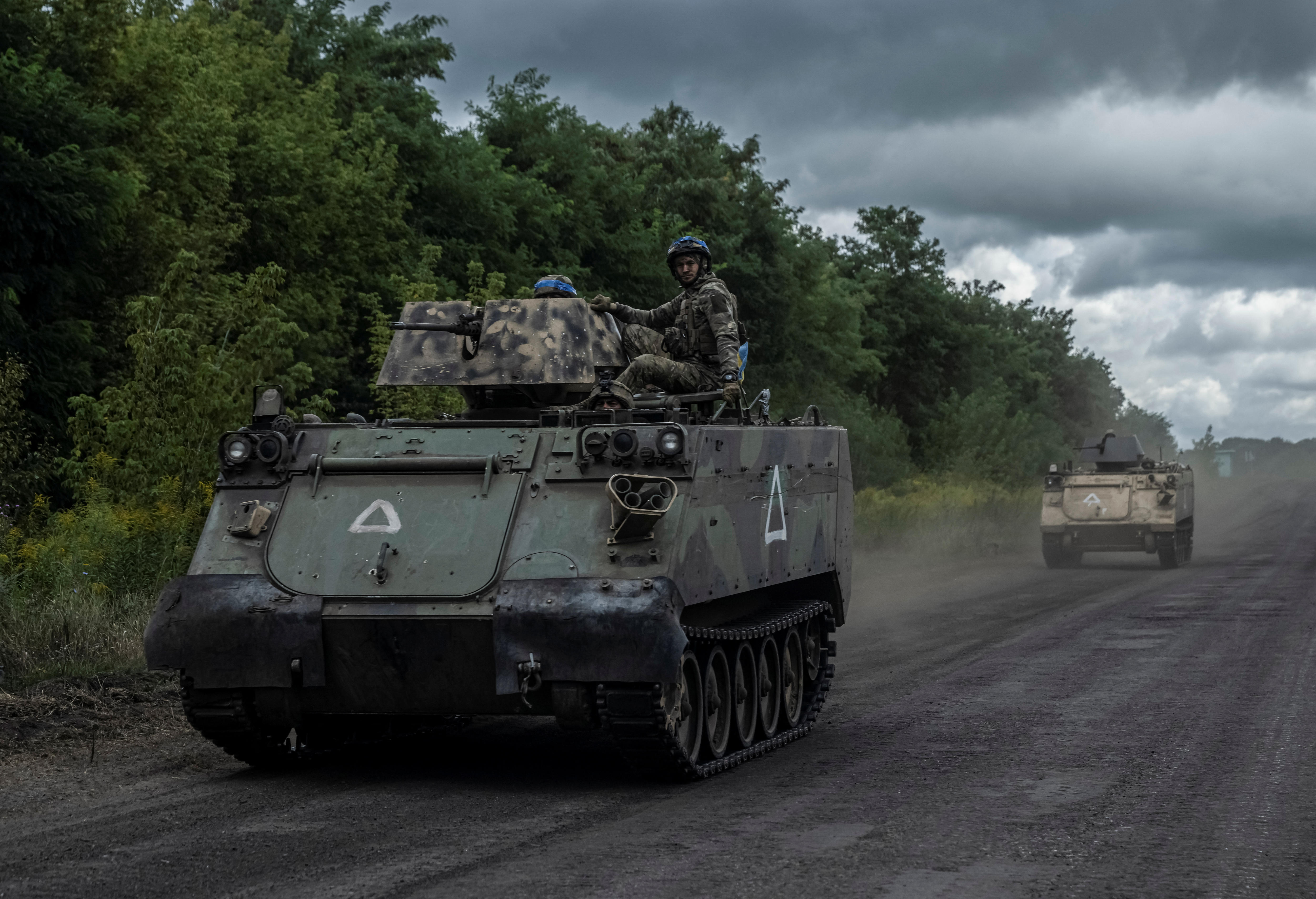Two soldiers sit on top of a small tank, driving in front of a second small tank, on a deserted road.