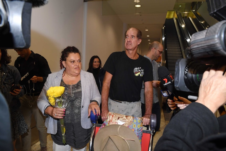 James Ricketson pushes a luggage cart, as a woman stands next to him with flowers, while at least two news cameras film