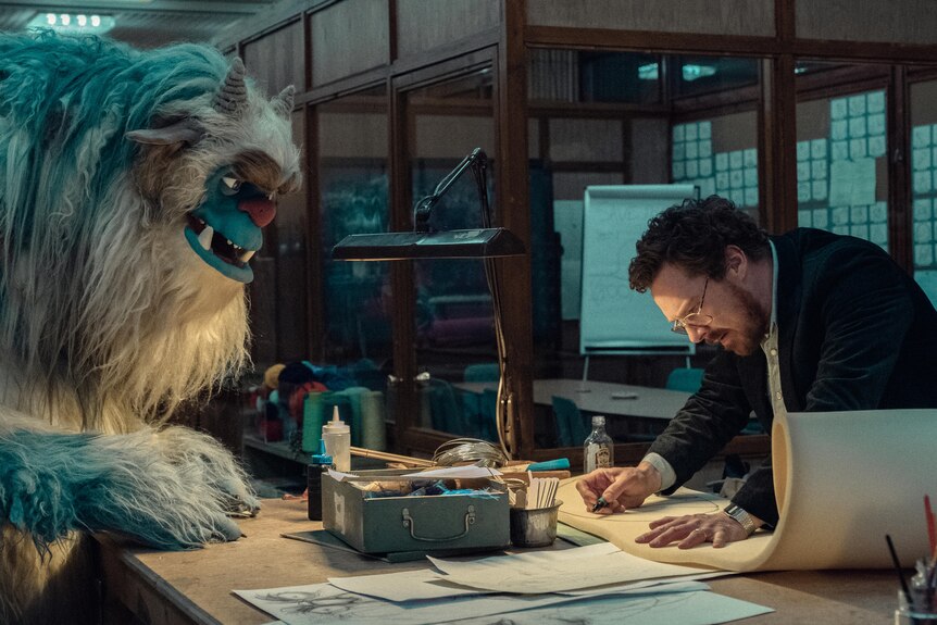 A giant blue monster puppet stands next to a man in a dark office