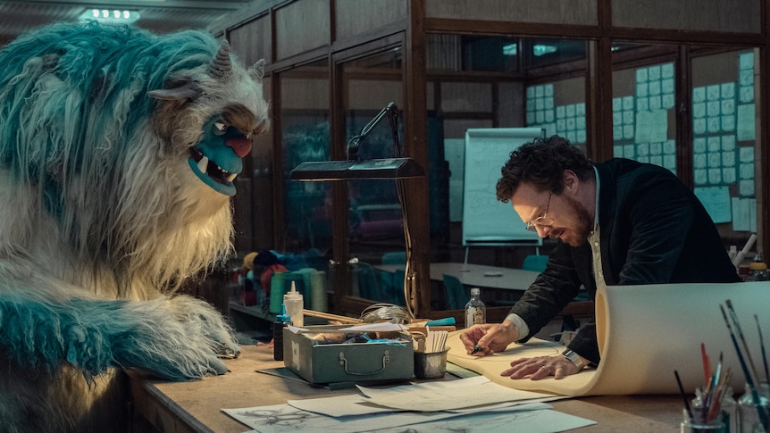 A giant blue monster puppet stands next to a man in a dark office