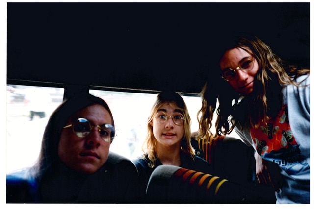 Three teenage boys sit on a bus. All three are wearing glasses, two seated while one stands