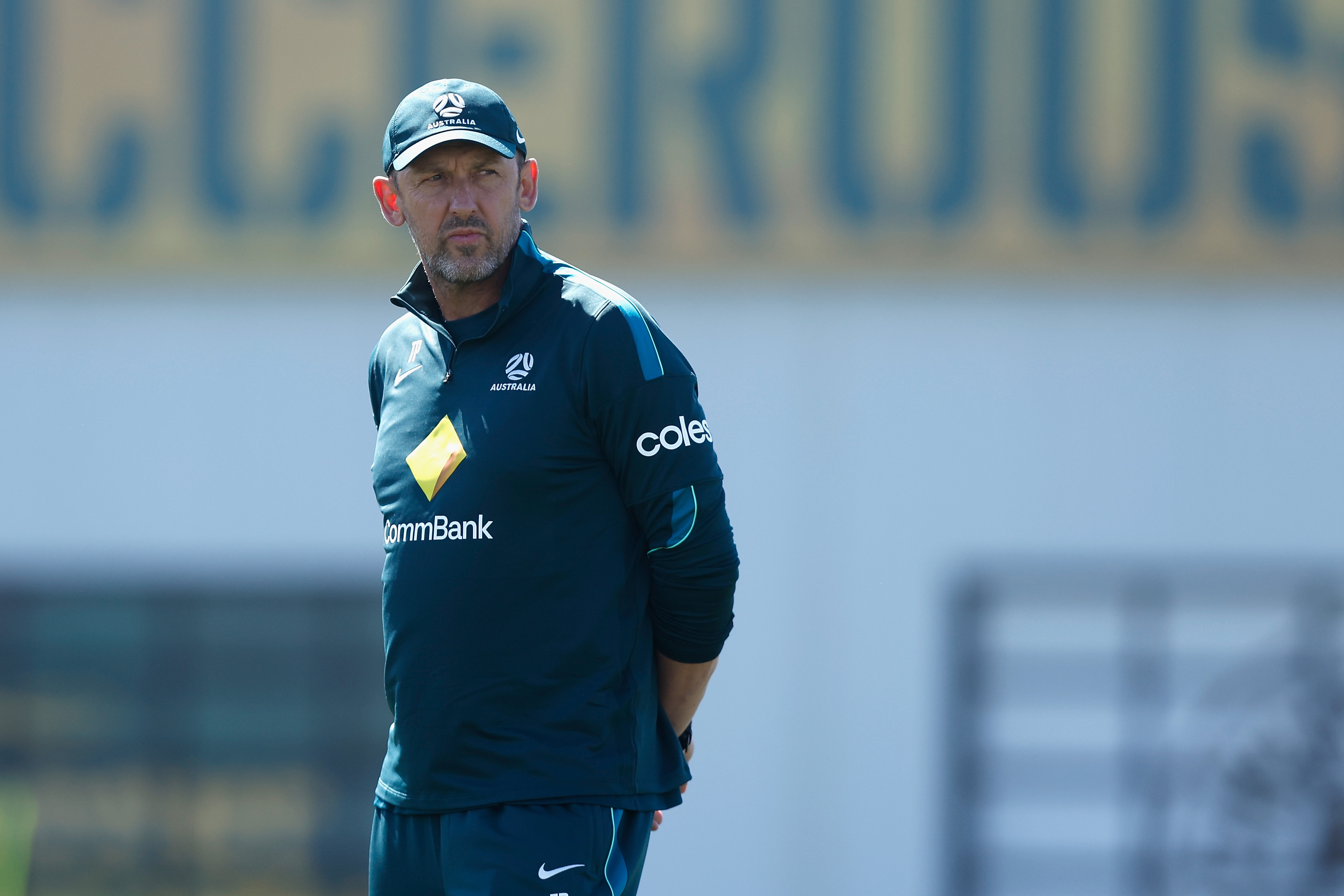 Tony Popovic looks over his shoulder at Socceroos training, wears green jersey cap with coles, CBA labels.