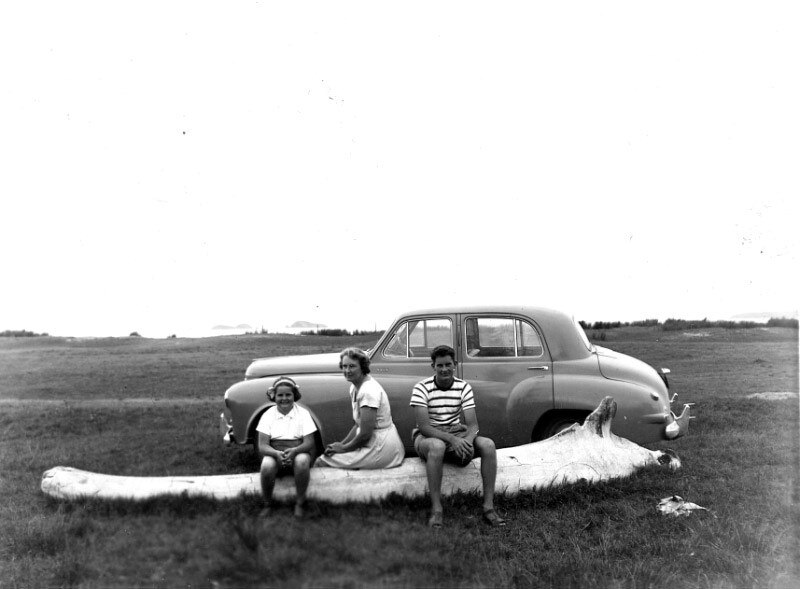 Black and white photo of one woman, a youth and a girl sitting on a whalebone in a vacant land, an old Holden is parked nearby.