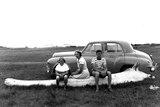 Black and white photo of one woman, a youth and a girl sitting on a whalebone in a vacant land, an old Holden is parked nearby.