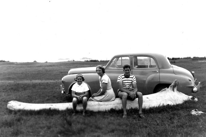 Black and white photo of one woman, a youth and a girl sitting on a whalebone in a vacant land, an old Holden is parked nearby.
