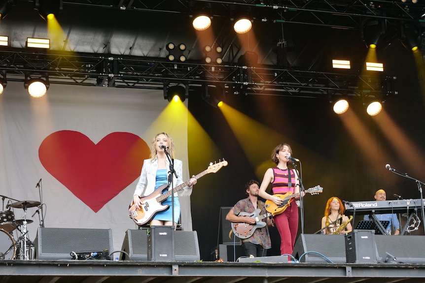 A band performing on a stage in front of a white banner with a red heart.