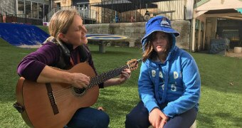 A lady in a purple jumper plays guitar with a disabled teenager wearing a blue jumper and matching bucket hat