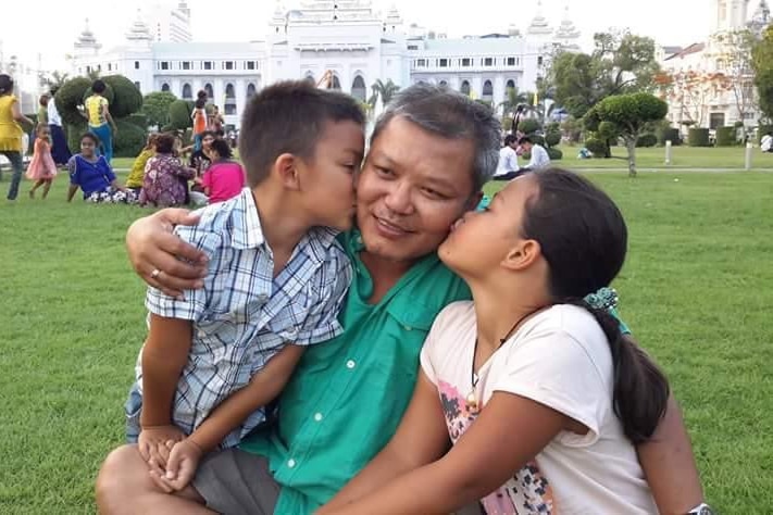Toe Zaw Latt pictured with his two children kissing him on the cheek as they sit on green grass.