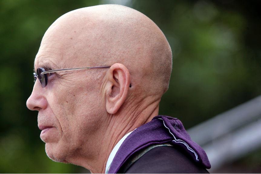 Profile of a bald man wearing glasses, and purple material around neck.