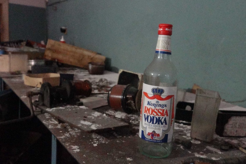 An old vodka bottle sits on a workbench inside an abandoned building.