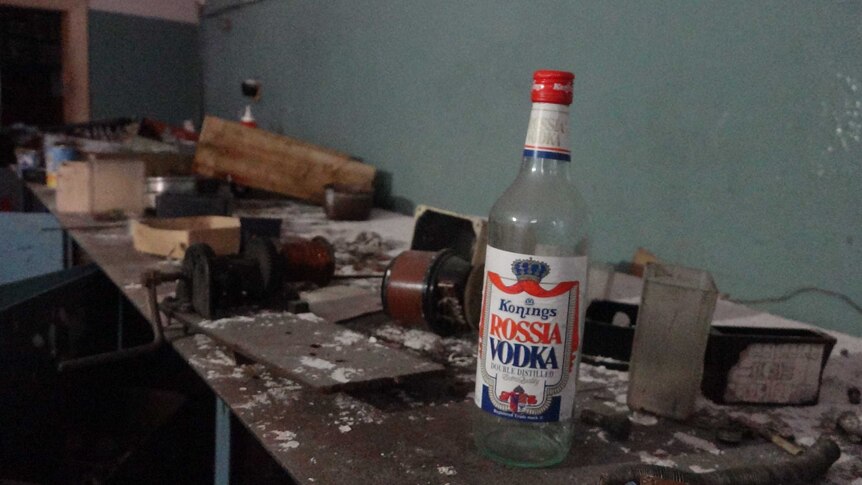 An old vodka bottle sits on a workbench inside an abandoned building.