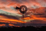 A windmill on a property is silhouetted against the red lights of the sunset reflecting off the underside of clouds.