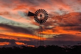 A windmill on a property is silhouetted against the red lights of the sunset reflecting off the underside of clouds.
