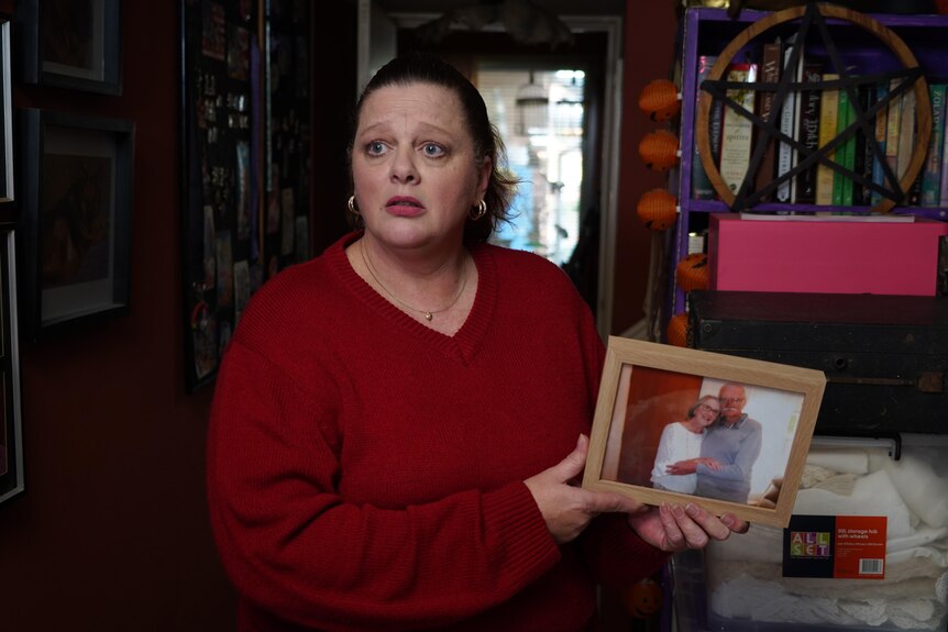 A woman looking sad wearing a red top holding a framed photo of an elderly couple