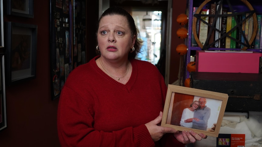 A woman looking sad wearing a red top holding a framed photo of an elderly couple