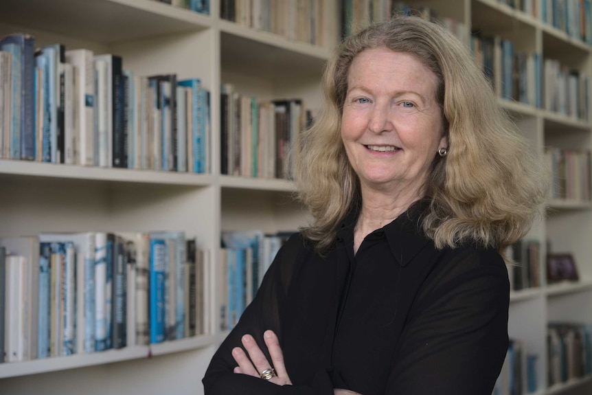 Judith Fox stands in front of book shelves, arms folded, smiling.