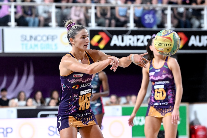A female netball player passes a ball hovering mid-way to the right of the frame.