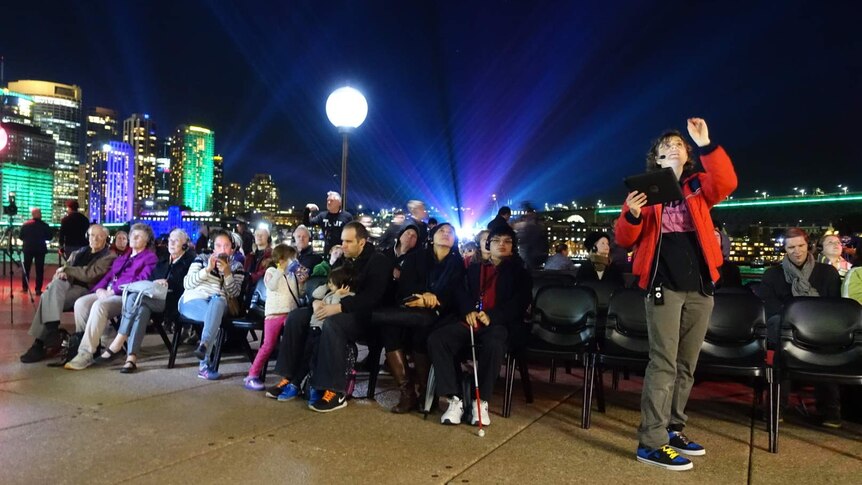 A number of vision impaired people sit listening to an audio description of the Vivid light show.