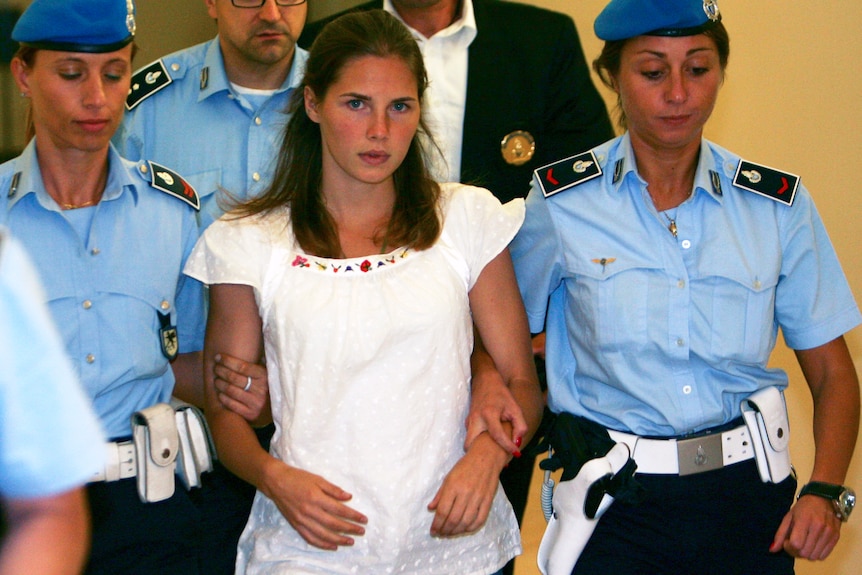 A young girl in a white peasant top is held by two police officers 