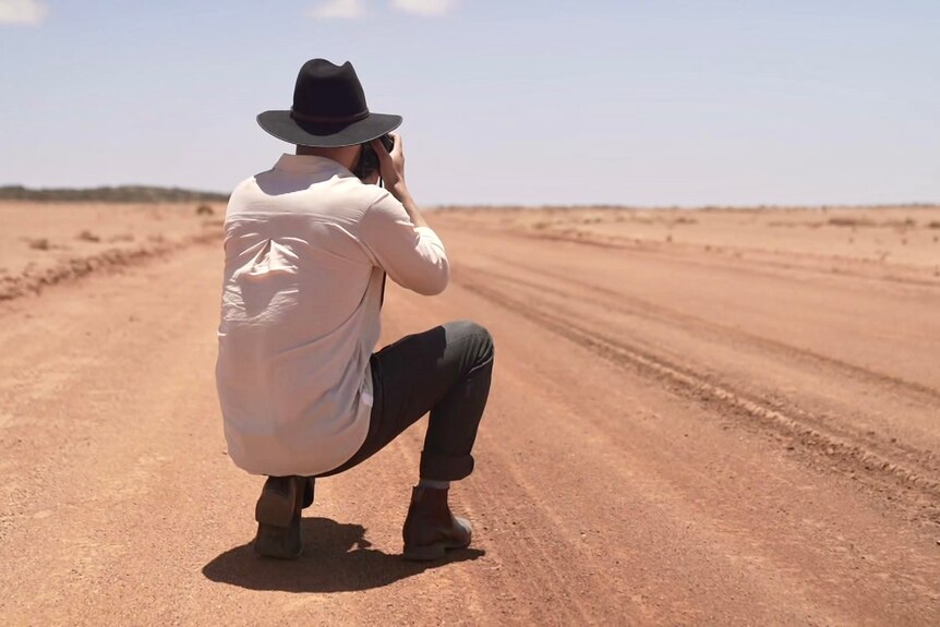 A man in a big black hat crouches down on a desert-looking road to take a photo.