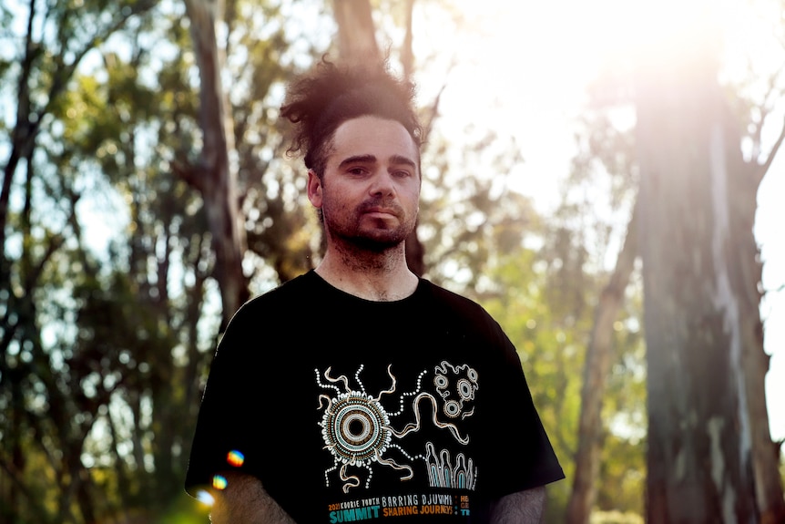 Man with black hair above head stands wearing black t-shirt smiling amid trees and sunlight