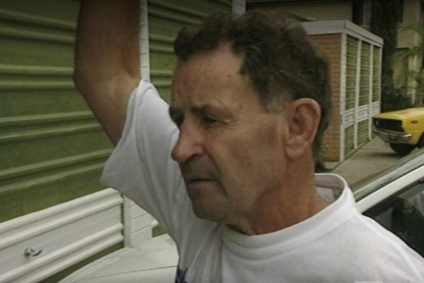A screen shot of John Coogan with his arm up wearing a white t-shirt and garage doors behind him