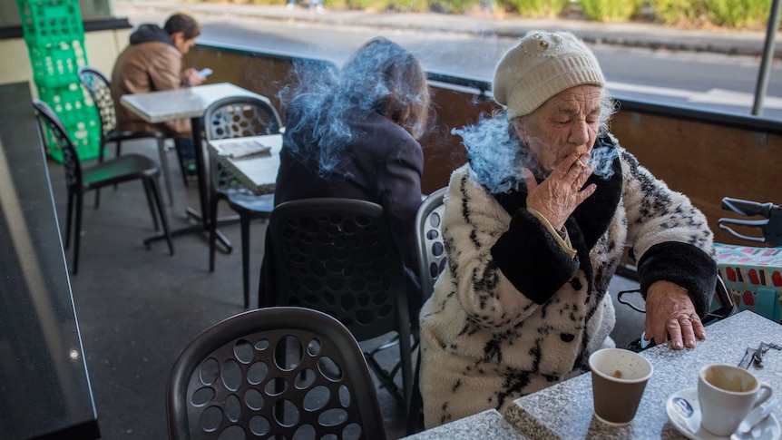 Bev Howlett smoking a cigarette at a table outdoors at a supermarket.