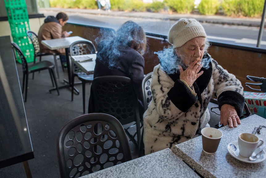 Bev Howlett smoking a cigarette at a table outdoors at a supermarket.