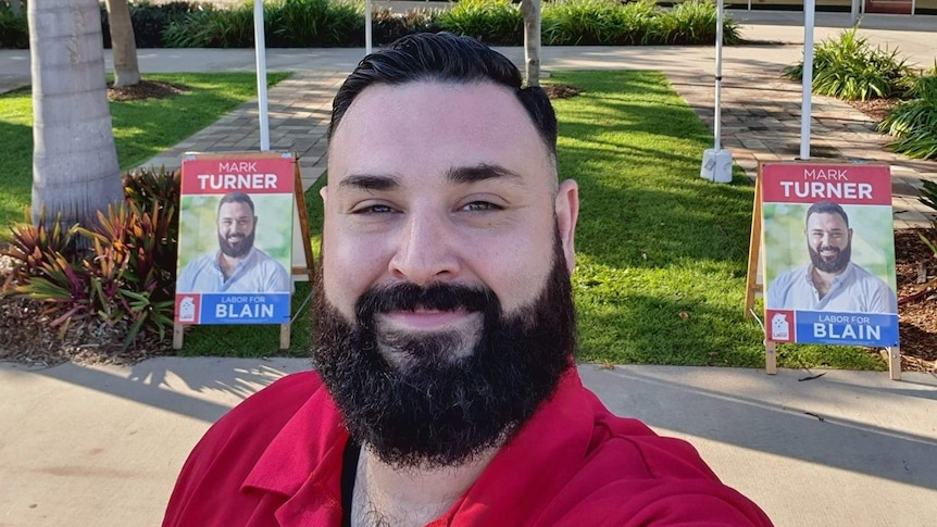 Politician Mark Turner smiles at the camera in front of two corflute A-frames with his name and photo.