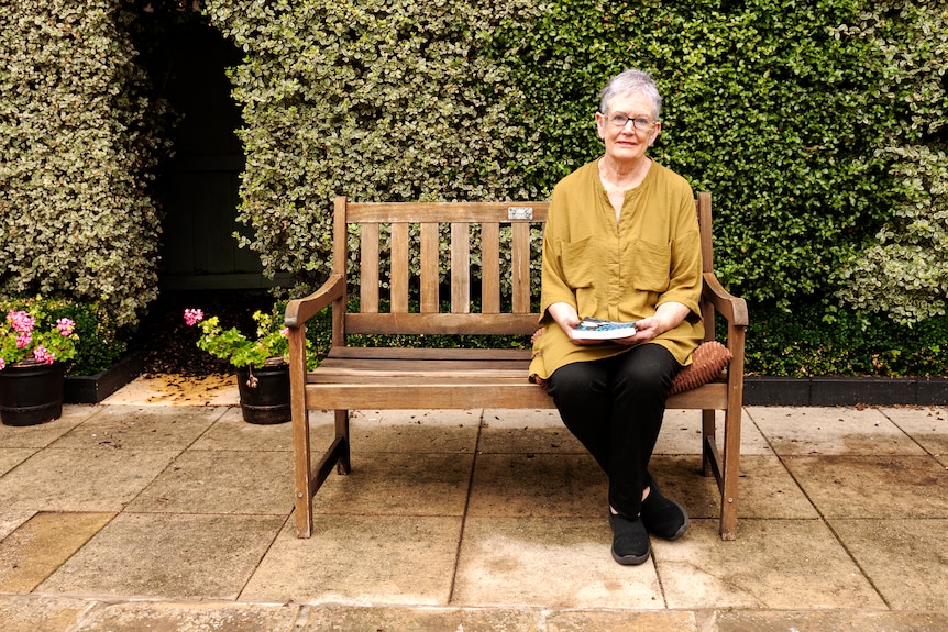 Woman wearing a yellow top sits on park bench with serious expression on her face. 