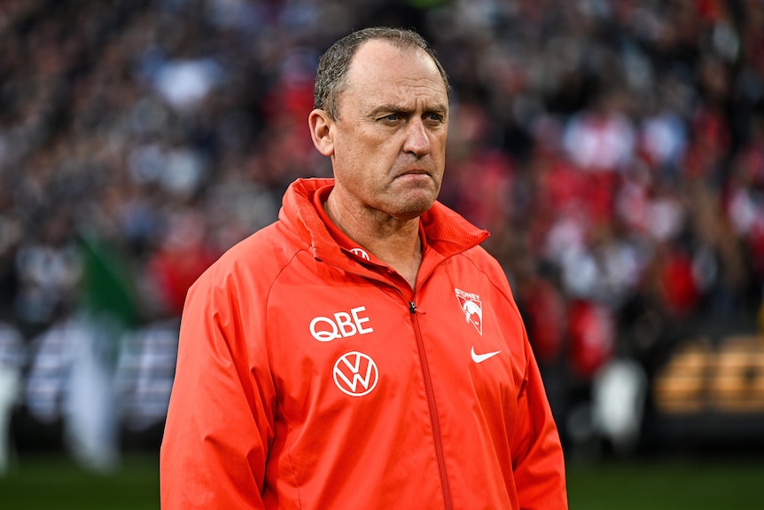 A dejected AFL coach clad in a red team jacket stands on the ground after a grand final loss.