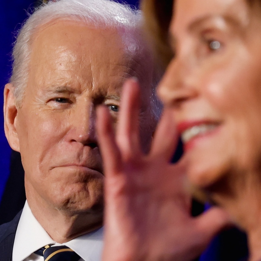 Joe Biden with a slight smile on his face watches Nancy Pelosi gesticulate 