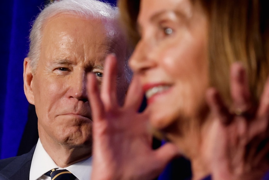Joe Biden with a slight smile on his face watches Nancy Pelosi gesticulate 