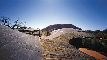 On a bright blue day, you view the undulating rooftop of a building with the top of Uluru poking out on the horizon.