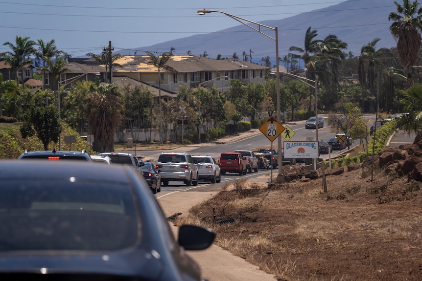 Cars are banked up on a road. Houses and mountains are seen in the background.