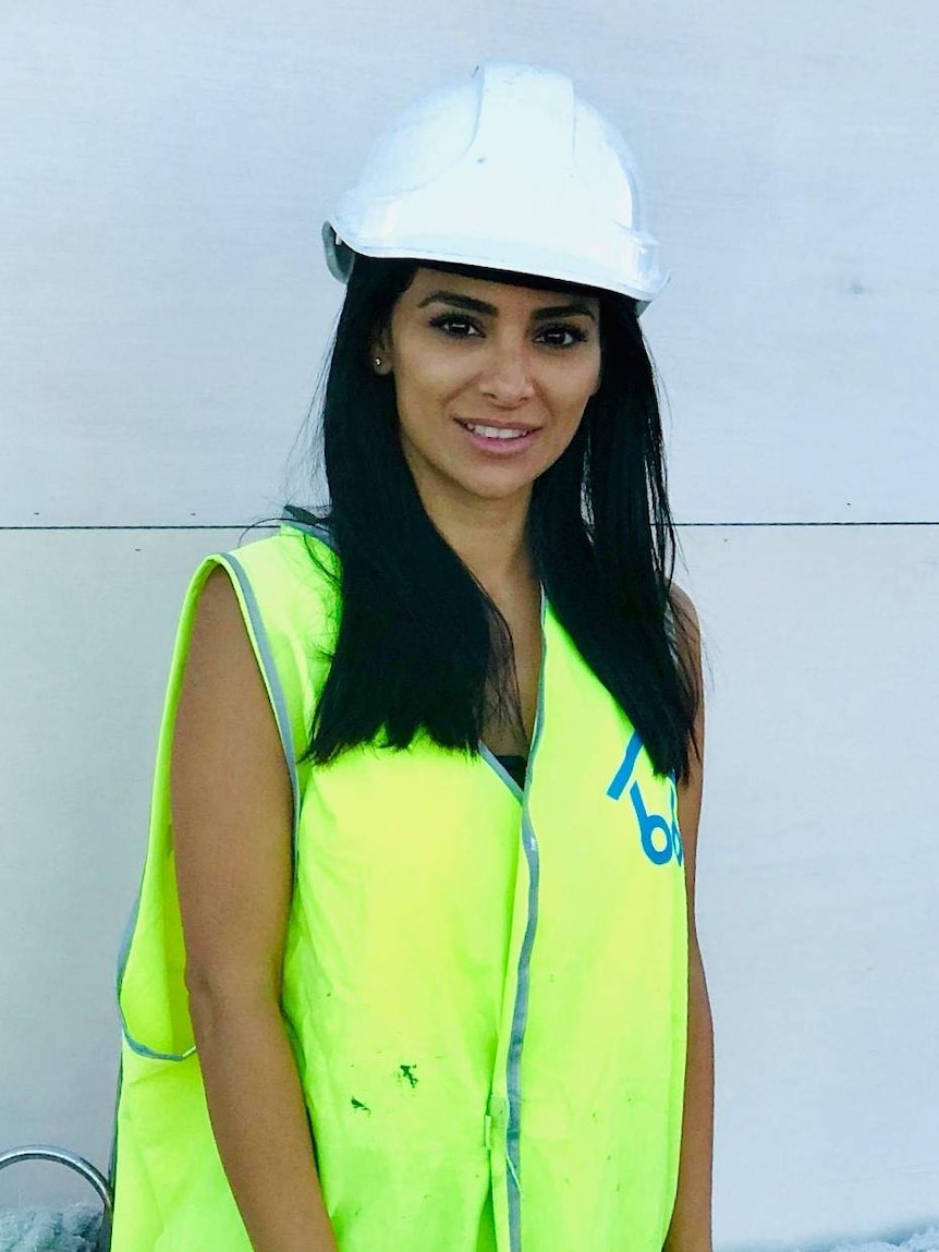 A woman stands looking at the camera. She is wearing a high-viz vest and a safety helmet.