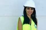 A woman stands looking at the camera. She is wearing a high-viz vest and a safety helmet.