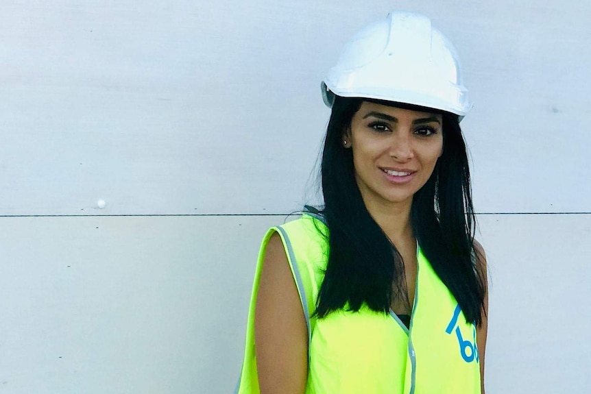 A woman stands looking at the camera. She is wearing a high-viz vest and a safety helmet.