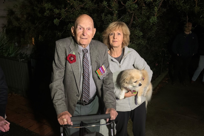 Fredrick Peirson, wearing a suit with a poppy and war medals, stands next to a woman holding a dog.