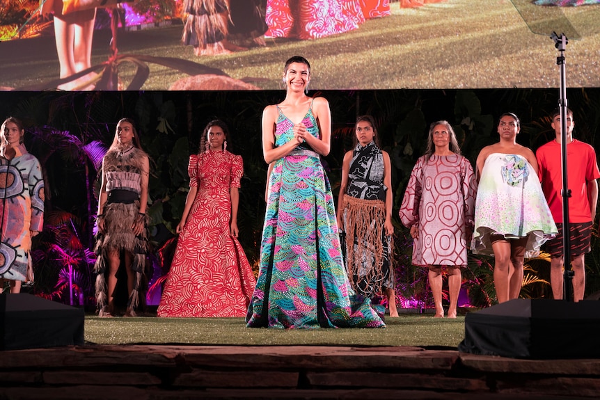 A group of fashion models stand in a line on the stage. A woman in a colourful dress stands in front of them.