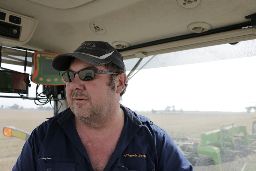 A man wearing a blue work shirt, sunglasses and a black cap sits in a tractor, with a field in the background.