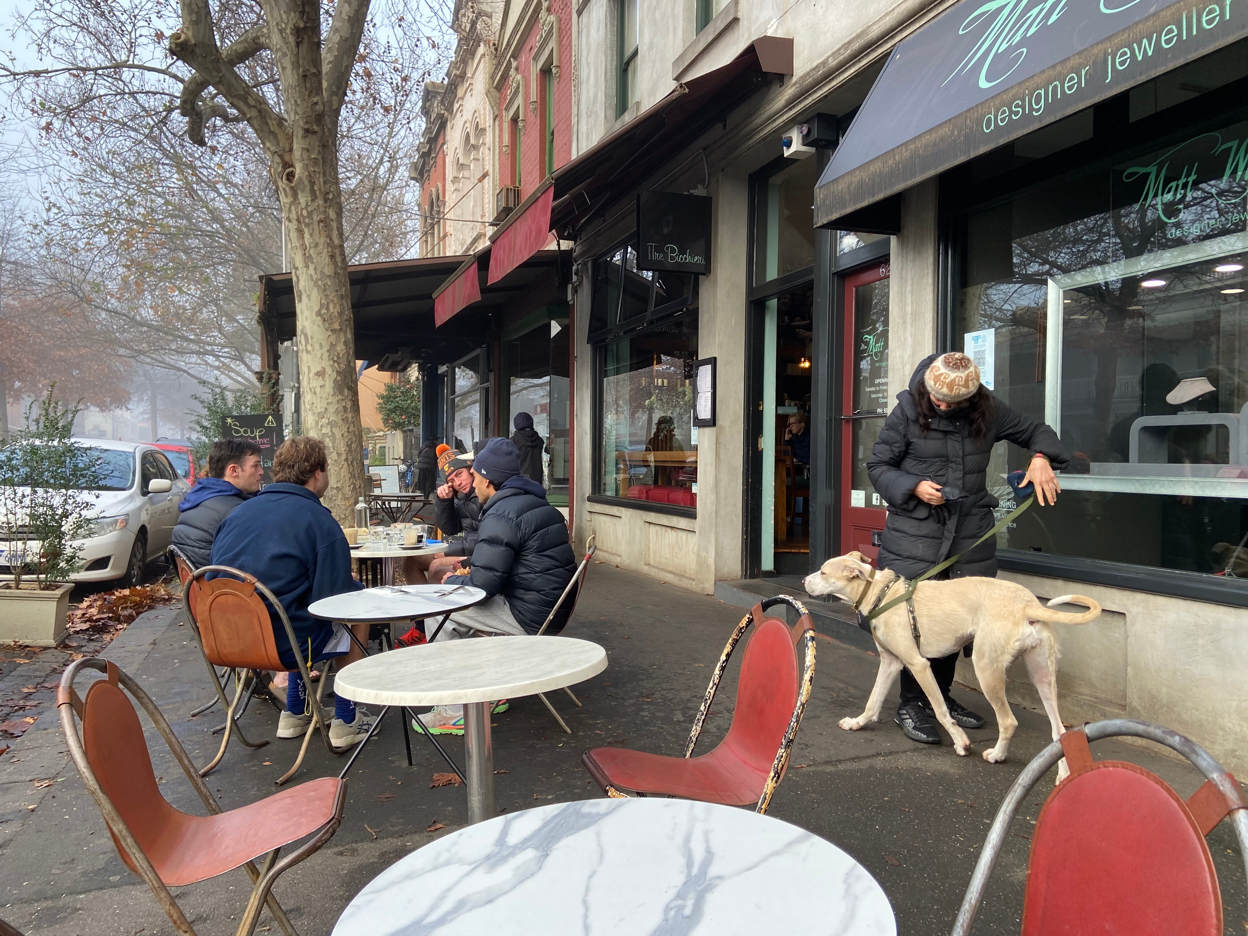 Four young men sit around an outdoor cafe table with coffees as a person walks their dog down the footpath.