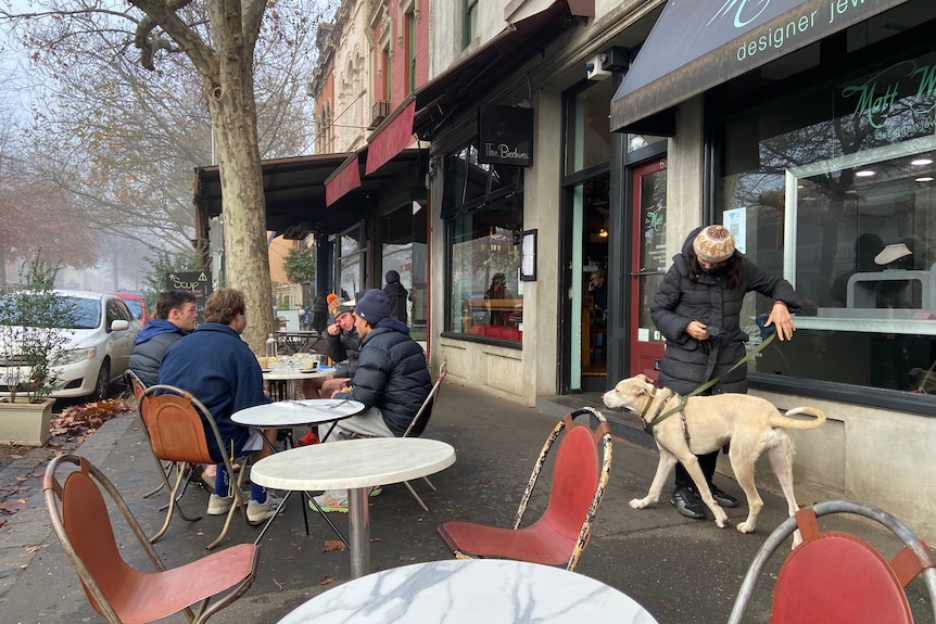 Four young men sit around an outdoor cafe table with coffees as a person walks their dog down the footpath.