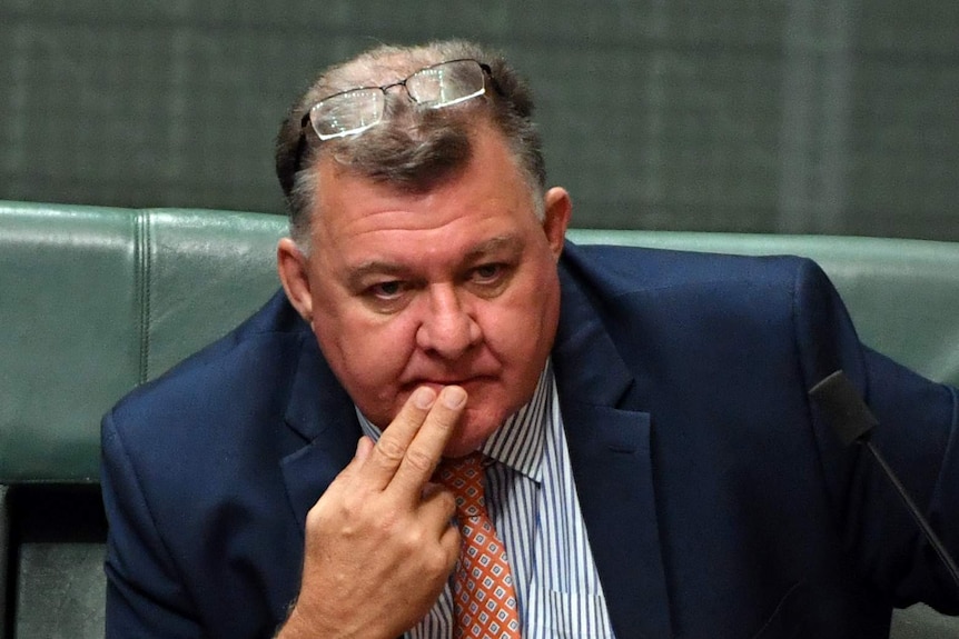 Craig Kelly leans forward in his seat in parliament, looking pensive with two fingers to his lips. His glasses sit atop his head