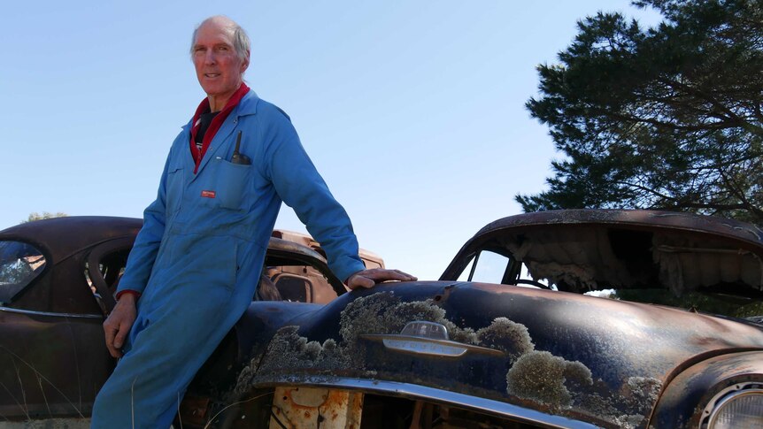 owner Wayne Flynn leans against rusty old car in a blue jumpsuit