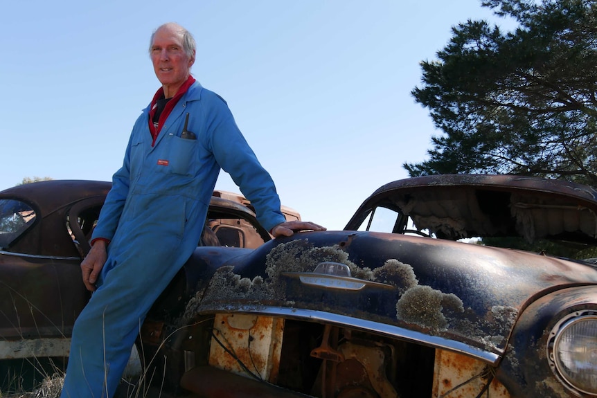 owner Wayne Flynn leans against rusty old car in a blue jumpsuit